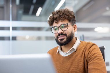 Gözlüklü ve sakallı genç profesyonel bir adam özgüvenle gülümsüyor, modern bir ofiste dizüstü bilgisayarla çalışıyor, özveriyi ve kurumsal başarıyı somutlaştırıyor.