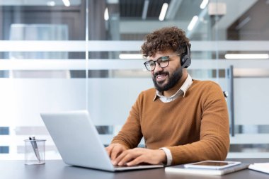 Bilgisayarda yazarken kulaklık ve gözlük takan, müşteri desteği sağlayan ya da modern ofis ortamından online iletişim sağlayan gülümseyen bir adam.