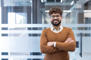 Sakallı ve gözlüklü genç profesyonel bir adam güvenle gülümsüyor, modern ofis ortamında kolları çapraz duruyor, başarıyı, uzmanlığı ve olumlu bir tutumu tasvir ediyor.