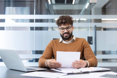 Modern bir ofis masasında mali raporları ve evrakları inceleyen gözlüklü genç işadamı bilgisayarındaki dosya, sözleşme ve verileri incelerken güvenle gülümsüyor.