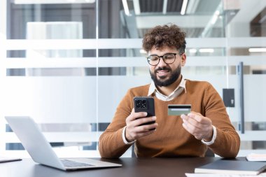 Elinde akıllı telefon ve kredi kartı olan genç bir adam ofis masasından güvenli bir şekilde online ödeme veya alışveriş yaparak modern e-ticaret ve dijital finansmanı kucaklıyor.