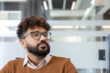 Kıvırcık saçlı ve sakallı genç bir adam ofis ortamında meditasyon yaparken sakin ve stresli bir an yaşıyor.