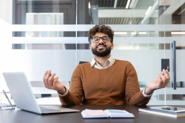 İş adamı masasında gözleri kapalı ve elleri meditasyon jesti içinde oturuyor, iç huzuru buluyor, farkındalığı yaşıyor ve mola sırasında stresi yönetiyor.