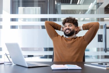 Mutlu gülümseyen, gözleri kapalı, elleri başının arkasında, tatmin olmuş ve başarılı hisseden, dizüstü bilgisayarındaki ve masasındaki görevlerini tamamladıktan sonra dinlenen bir adam.