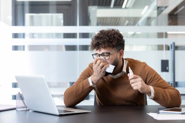Genç işadamı soğuk algınlığı ya da alerjiden muzdarip, modern ofis ortamında masasında çalışırken burnunu mendille silip burun spreyi tutuyor.