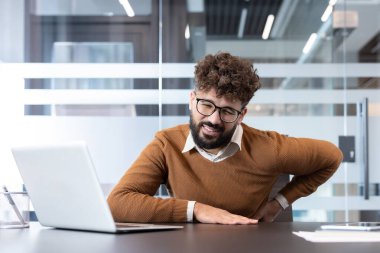 Sırt ağrısı ya da bel ağrısı çeken genç bir adam, ofiste dizüstü bilgisayarı üzerinde çalışırken sabit bir yaşam tarzı ve zayıf duruşu nedeniyle rahatsızlık ve gerginlik hissediyor.