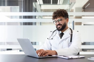 Genç bir erkek doktor, steteskobu boynunda, dizüstü bilgisayarla yazarken gülümsüyor ve modern bir klinikten tele-sağlık danışmanlığı hizmeti veriyor.