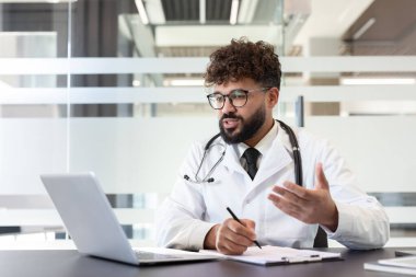 Laboratuvar önlüğü giyen ve steteskop kullanan erkek doktor bilgisayardaki bir hastayla çevrimiçi video konferansı yapıyor.