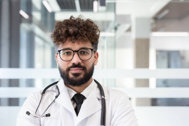 Sakallı, kıvırcık saçlı, gözlüklü, steteskop takan ve beyaz önlüklü genç bir erkek doktor modern bir klinikte ya da hastane koridorunda güvenle duruyor.