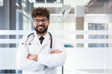 Beyaz laboratuvar önlüğü ve steteskop giyen genç bir erkek doktor, kollarını kavuşturmuş sıcak bir şekilde gülümsüyor, modern bir hastane ya da klinik ortamında güvenle duruyor.