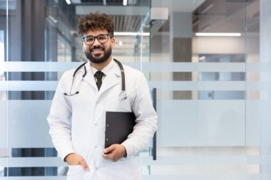 Genç doktor gülümsüyor, laboratuvar önlüğü ve steteskop giyiyor, elinde bir pano tutuyor, çağdaş bir klinikte ya da hastane lobisinde duruyor, tıbbi profesyonelliği ve sağlık hizmetlerini temsil ediyor.