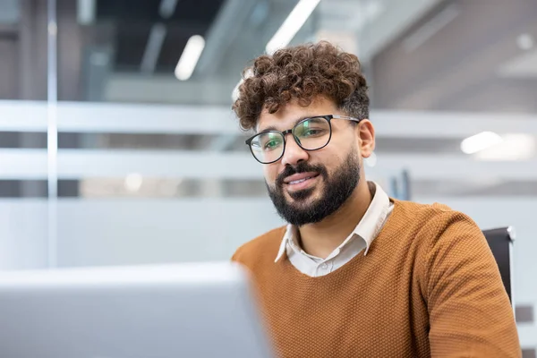 Gözlüklü ve sakallı genç profesyonel bir adam özgüvenle gülümsüyor, modern bir ofiste dizüstü bilgisayarla çalışıyor, özveriyi ve kurumsal başarıyı somutlaştırıyor.