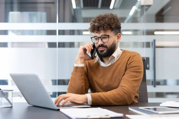 Genç sakallı girişimci, gözlüklü, aktif olarak bilgisayarda yazarken bir telefon görüşmesi yapan, modern ofis masasından birden fazla işi yöneten.
