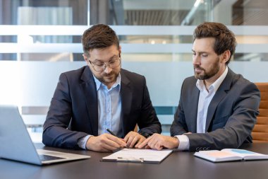 Konferans masasında takım elbiseli iki işadamı bir sözleşmeyi gözden geçiriyorlar. Biri şartlara işaret ederken diğeri de imzalamaya hazırlanıyor. Profesyonel bir ofiste resmi bir ortaklık için pazarlık yapıyorlar.