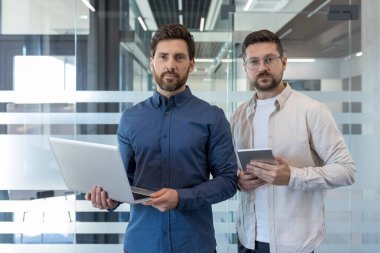 Bir ofiste duran iki kendine güvenen iş adamı, biri dizüstü bilgisayar ve diğeri tablet, takım çalışmasını, teknolojiyi ve modern bir şirket ortamını temsil ediyor.