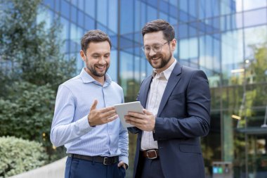 İki gülümseyen işadamı dijital tablete bakıyor, kurumsal stratejiyi tartışıyor ve arka planda modern cam bir bina olan şehir ortamında birlikte çalışıyorlar.