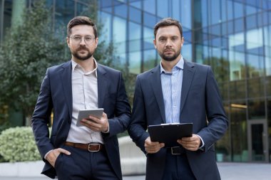İki kendine güvenen iş ortağı, biri dijital tabletli diğeri ise şirket takım çalışmasını ve başarısını simgeleyen bir pano ile modern cam ofis binasının önünde duruyor.