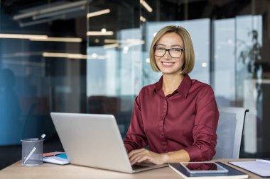 Profesyonel bir iş kadını masasında otururken kameraya gülümser, dizüstü bilgisayarda yazar ve başarılı bir kariyer ve kurumsal çevreyi yansıtır.