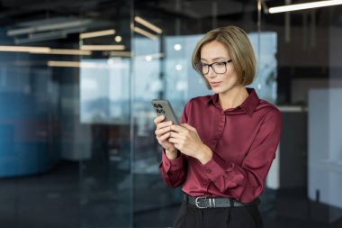 Çağdaş bir ofis yerinde duran gözlüklü bir iş kadını, akıllı telefonuna odaklanmış, dijital iletişimle meşgul ve mesleki görevlerini yerine getiriyor.