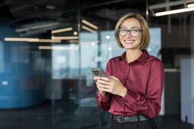 Genç iş kadını gülümsüyor, gözlük takıyor ve modern bir ofiste cep telefonu tutuyor. Profesyonel bir ortamda iletişim, teknoloji ve başarıyı temsil ediyor.