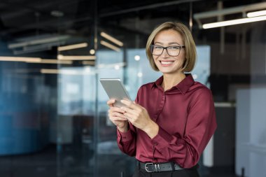 Gözlüklü ve bordo gömlekli gülümseyen iş kadını, elinde tablet, yarım vücut portresi, profesyonellik ve dijital takım çalışması taşıyan modern bir ofiste güvenle duruyor.