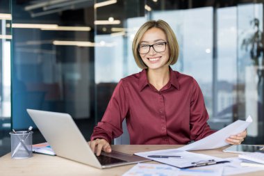 Modern ofisteki iş kadını masada otururken kameraya gülümsüyor, dizüstü bilgisayar üzerinde çalışıyor ve elinde önemli belgeler tutuyor, kendinden emin ve profesyonel girişimci