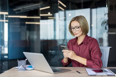 Günümüz kurumsal ortamında, elinde laptop ve belgelerle ahşap bir masada otururken akıllı telefonuyla iletişimi sağlayan gözlüklü bir iş kadını.