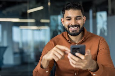 Hintli işadamı gülümsüyor, iletişimden keyif alıyor, modern bir ofiste cep telefonu tutuyor, mesaj yazıyor veya sosyal medyayı kontrol ediyor, iletişim ve teknolojiyi temsil ediyor.