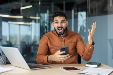 İşadamı şok ve hüsran içinde akıllı telefonuna bakıyor, ağzı sarkık ve ofis masasındaki kötü haberlere tepki verirken elini uzatıyor, stresli ve şaşkın bir şekilde.