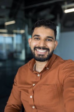 Sakallı gülümseyen Hintli işadamı mutlu ve olumlu bir ifade sergiliyor, modern bir ofiste ya da ortak çalışma alanında güvenle duruyor, başarı ve profesyonelliği temsil ediyor.