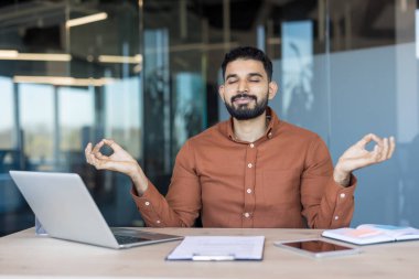 Genç Hintli iş adamı modern masasında farkındalık uyguluyor, gözleri kapalı ve elleri Mudra 'da odaklanmış bir iş günü molası sırasında sakin ve stresli bir şekilde dinleniyor.