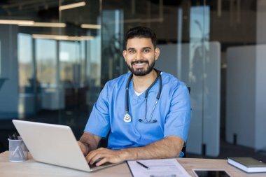 Modern ofiste steteskoplu Hintli doktor bilgisayarına gülümsüyor, tele sağlık danışmanlığı ve dijital sağlık desteği sunuyor, kendinden emin ve profesyonel.