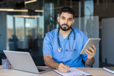 Mavi önlüklü ve steteskoplu erkek doktor tablet ve dizüstü bilgisayarla masada oturuyor, modern teletıp, dijital dokümantasyon ve klinik bakım ile ilgili notlar odaklı portre yazıyor.