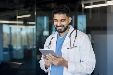 Beyaz önlük ve steteskop giyen Hintli doktor, modern bir hastane veya klinik ortamında dijital tablet kullanırken gülümsüyor, tıbbi yenilik ve kendine güvenen sağlık hizmetlerini temsil ediyor.