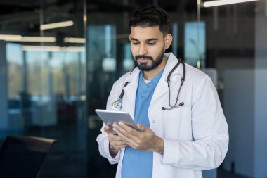 Beyaz önlüklü ve steteskoplu genç erkek doktor, modern bir klinikteki hasta verilerini veya sağlık bilgilerini inceleyen dijital bir tablete bakıyor.
