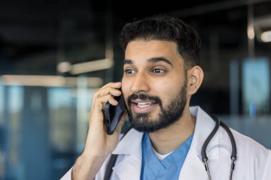 Klinikte sakallı ve steteskoplu genç bir erkek doktor yoğun bir teletıp muayenesi sırasında hasta bakımını ve tıbbi tavsiyeleri tartışan bir telefona odaklanmış.