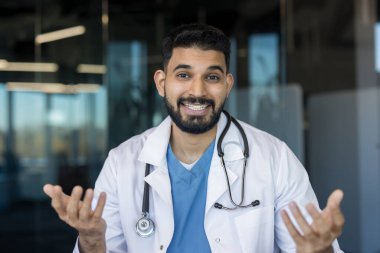 Laboratuvar önlüğü ve steteskop giymiş Hintli erkek doktor kameraya gülümsüyor, tıbbi bir konsepti açıklıyor veya online danışmanlık veya tele sağlık toplantısı sırasında tavsiye veriyor