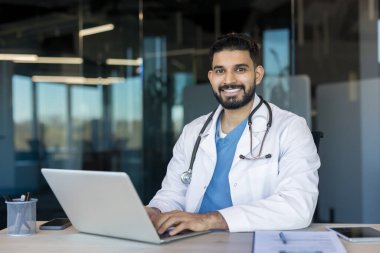 Genç Hintli erkek doktor beyaz önlük ve steteskop giyiyor, bir ofis masasında dizüstü bilgisayarla çalışırken gülümsüyor, modern sağlık, tele sağlık ve ulaşılabilir tıbbi muayeneleri sembolize ediyor.