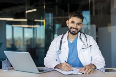 Beyaz önlüklü genç Hintli erkek doktor masaya gülümsüyor, laptopun ve tabletin yanındaki bir panoya yazıyor, modern teletıp ve profesyonel bakımı iletiyor.