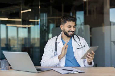 Üniformalı ve steteskoplu mutlu erkek doktor dijital tabletle video görüşmesi yapıyor. Masasında dizüstü bilgisayarı olan modern bir ofisten uzaktan tıbbi danışmanlık ve çevrimiçi sağlık hizmeti sunuyor.