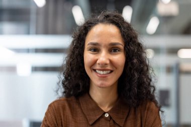 Genç kıvırcık saçlı iş kadını modern ofis fotoğrafında güvenle gülümsüyor, pozitifliğini, profesyonelliğini ve başarılı liderliğini gündelik bir şirket ortamında aktarıyor.