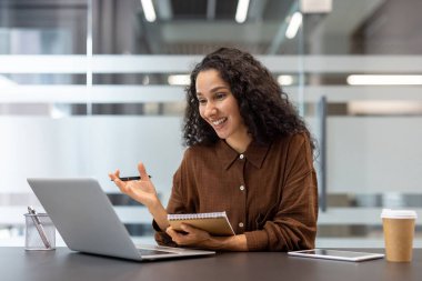 Modern bir ofisten dizüstü bilgisayarla çevrimiçi bir video görüşmesine katılan mutlu genç kadın notlar alıyor ve iş kavramlarını tartışıyor.