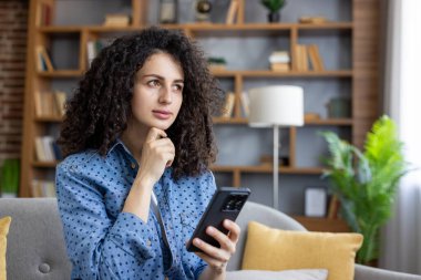 Genç bir kadın akıllı telefonu evdeki bir kanepede tutarken düşünüyor, yansıtıyor, tarıyor ve çevrimiçi bağlanıyor, dijital iletişimi ve tefekkür etmeyi temsil ediyor.