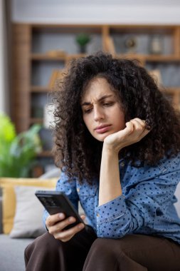 Kıvırcık saçlı, üzgün ve üzgün bir genç kadın evdeki cep telefonunda üzücü bir mesaj ya da kötü haber okurken hayal kırıklığı ve hayal kırıklığı yaşıyor.