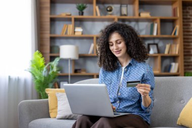 Evdeki kanepede gülümseyen genç bir kadın, elinde bir kredi kartı ve internetten alışveriş yapmak için dizüstü bilgisayar kullanıyor ve e-ticaret ödemesini tamamlıyor, uygun dijital alışverişin tadını çıkarıyor.