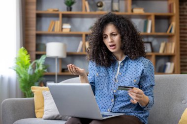 Evde bir kanepede oturan hayal kırıklığına uğramış genç bir kadın, online alışveriş veya bankacılık ile ilgili sorunlar yaşıyor, kredi kartını tutarken kafası karışmış görünüyor ve dizüstü bilgisayar kullanıyor.