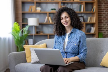 Genç bir kadın dizüstü bilgisayarda gülümsüyor ve daktilo kullanıyor. Oturma odasındaki kanepede rahat bir şekilde çalışıyor ya da çalışıyor. Arkasında bir kitaplık ve yeşil bir bitki var.
