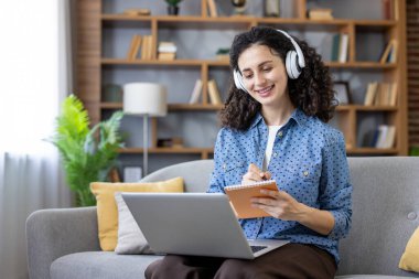 Kulaklık takan, gülümseyen ve notlar alan bir kadın modern oturma odasındaki bir kanepede çalışırken, e-öğrenme ve evdeki uzaktan çalışmaya odaklandı.