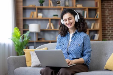 Kablosuz kulaklık takan, gülümseyen, gri bir koltukta rahat bir şekilde oturan ve dizüstü bilgisayarda yazan, online eğitim alan ya da evden çalışan bir kadın.