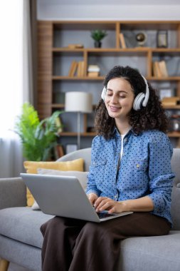 Kanepede oturan kıvırcık saçlı bir kadın, kulaklık takan ve dizüstü bilgisayar kullanan, çalışırken gülümseyen ya da rahat bir ev ortamında uzaktan çalışan.